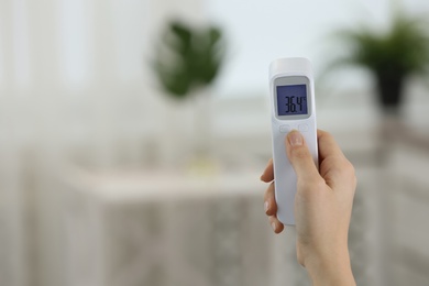 Closeup view of woman with infrared thermometer on blurred background, space for text. Checking temperature during Covid-19 pandemic