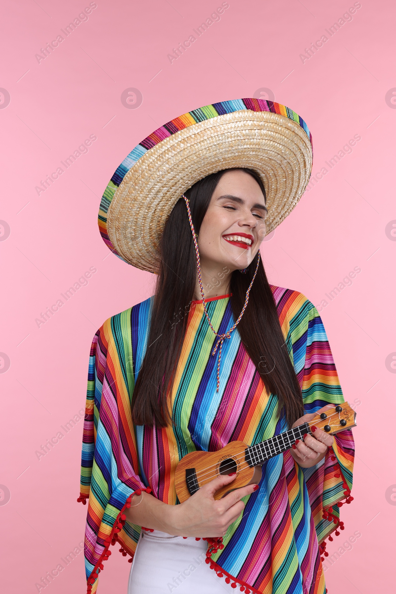 Photo of Young woman in Mexican sombrero hat and poncho playing ukulele on pink background