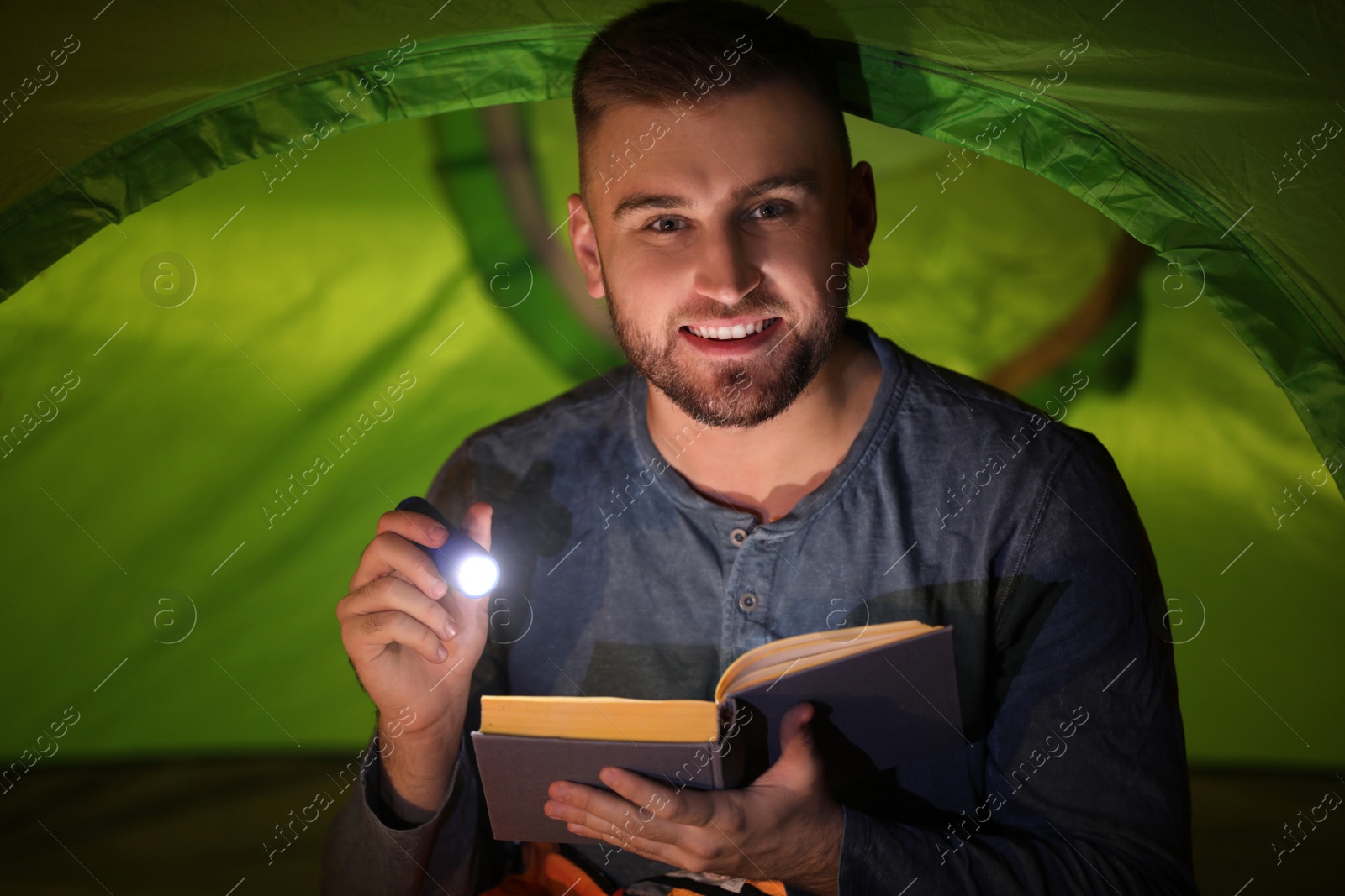 Photo of Young man with flashlight reading book in tent