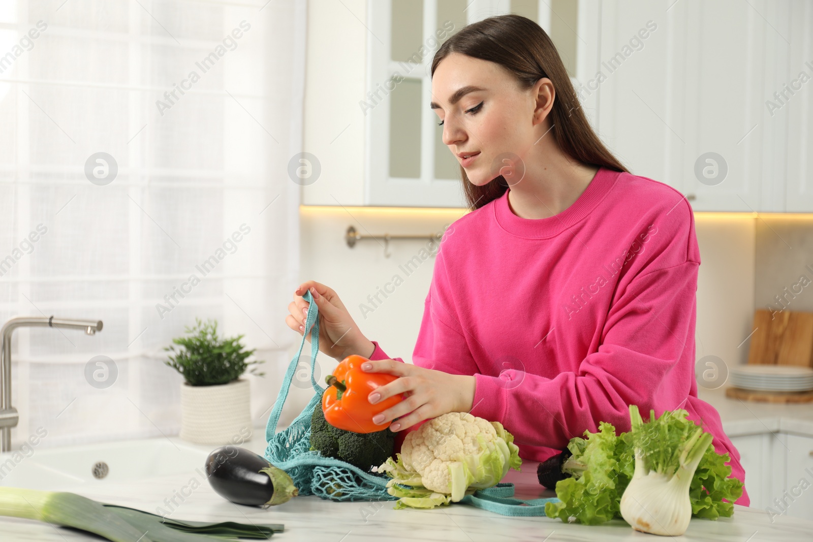 Photo of Woman taking pepper out from string bag at light marble table in kitchen