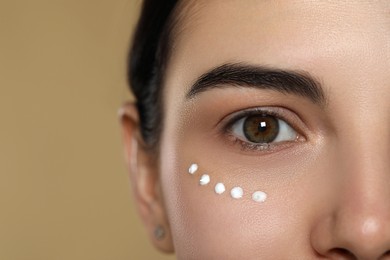 Photo of Young woman with cream on skin under eye against beige background, closeup