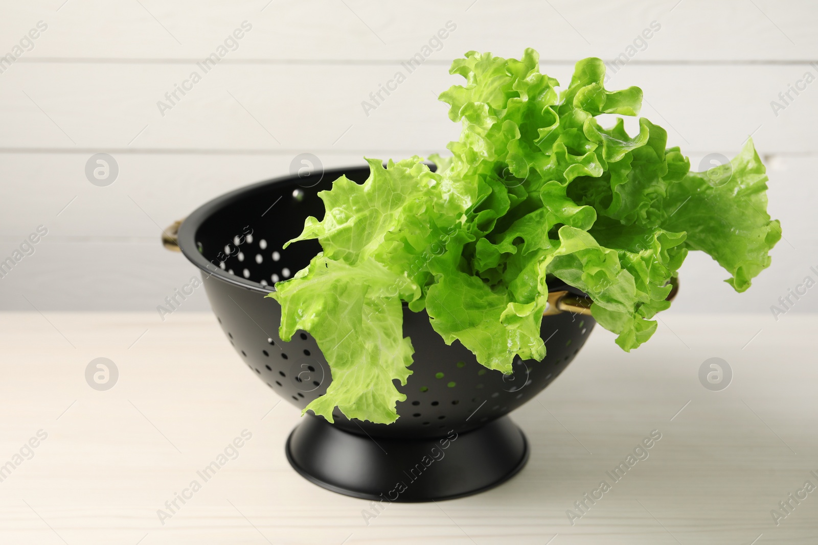 Photo of Fresh lettuce in black colander on white wooden table