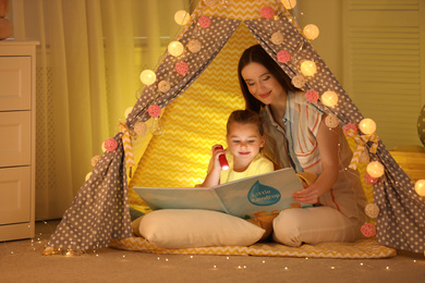 Mother and daughter with flashlight reading book in play tent at home