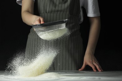 Photo of Woman sieving flour at table against black background, closeup