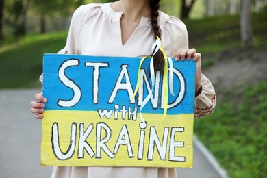 Photo of Young woman holding poster in colorsnational flag and words Stand with Ukraine outdoors, closeup