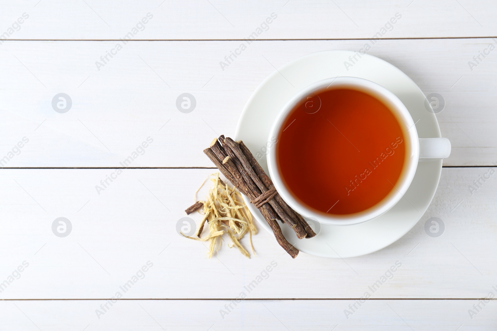 Photo of Aromatic licorice tea in cup and dried sticks of licorice root on white wooden table, top view. Space for text