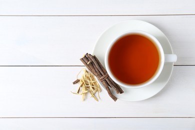Aromatic licorice tea in cup and dried sticks of licorice root on white wooden table, top view. Space for text