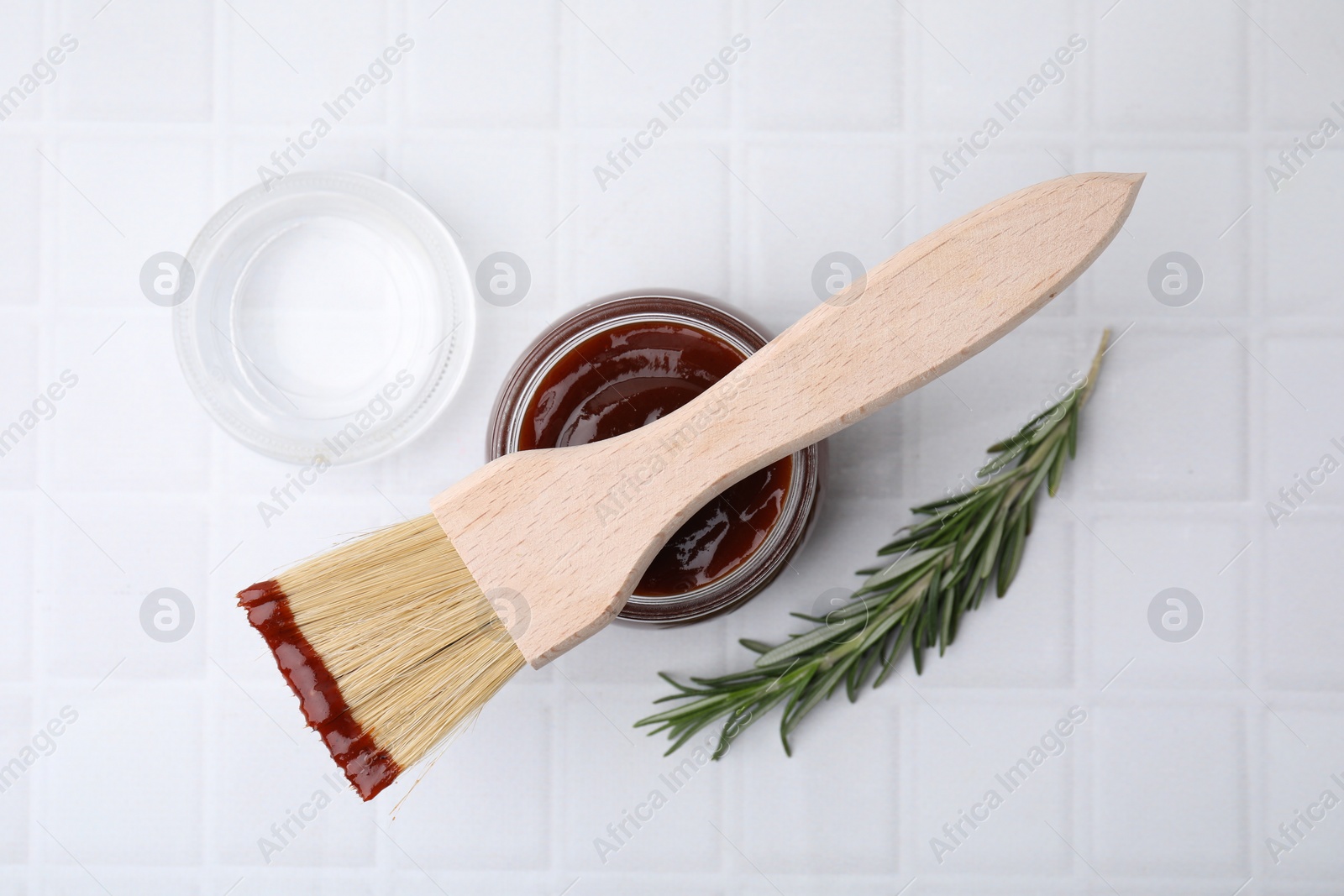 Photo of Marinade in jar, rosemary and basting brush on white tiled table, flat lay