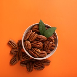 Photo of Bowl with tasty pecan nuts and green leaves on orange background, flat lay