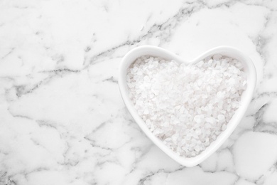 Photo of Heart shaped bowl of white sea salt on marble table, top view with space for text. Spa treatment