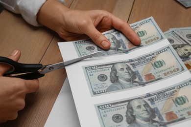 Photo of Counterfeiter cutting dollar banknotes with scissors at wooden table, closeup. Fake money concept