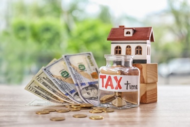 Photo of Glass jar with label "TAX TIME", money and model of house on wooden table