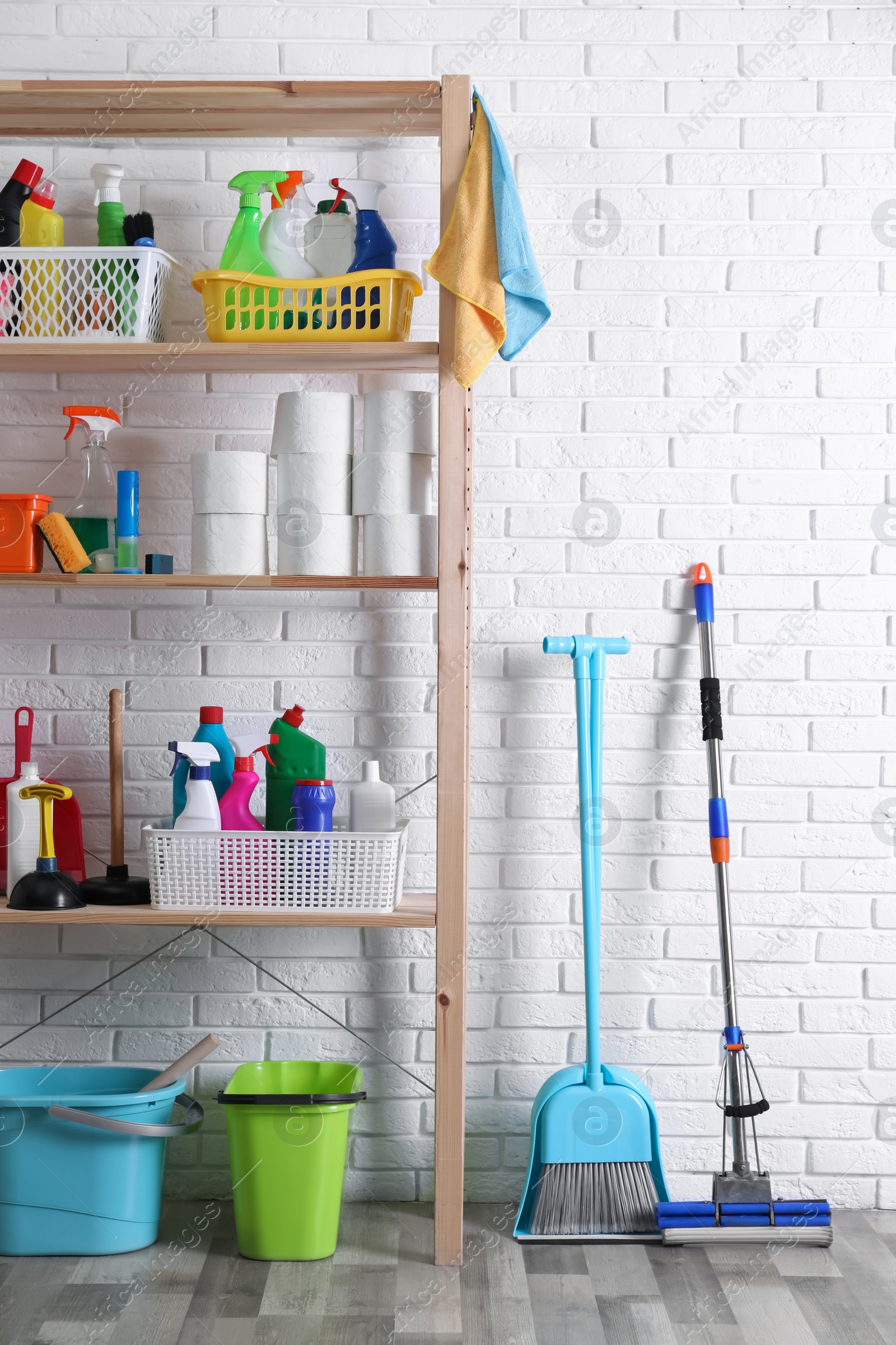 Photo of Shelving unit with detergents, cleaning tools and toilet paper near white brick wall indoors