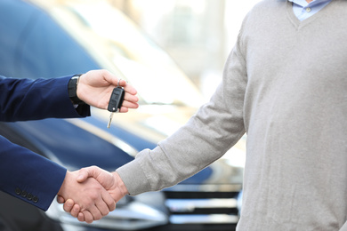 Photo of Salesman giving key to customer while shaking hands in modern auto dealership, closeup. Buying new car