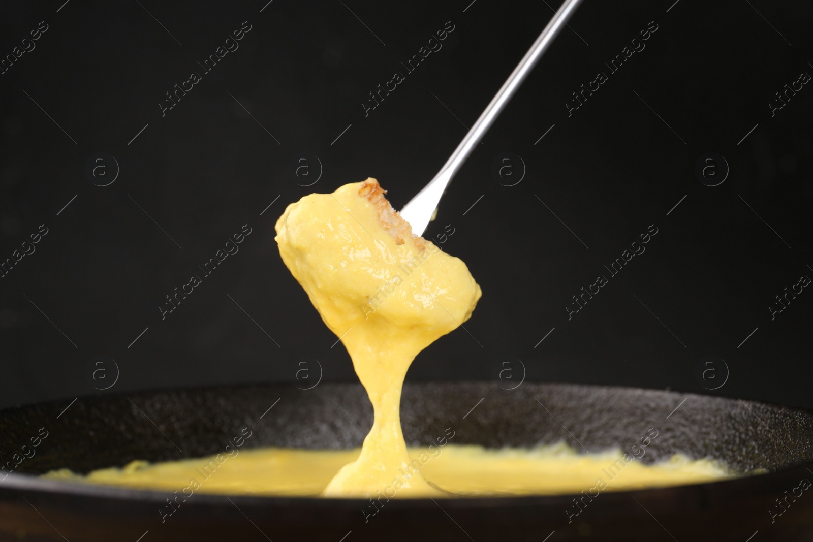 Photo of Dipping piece of bread into fondue pot with tasty melted cheese against dark gray background, closeup