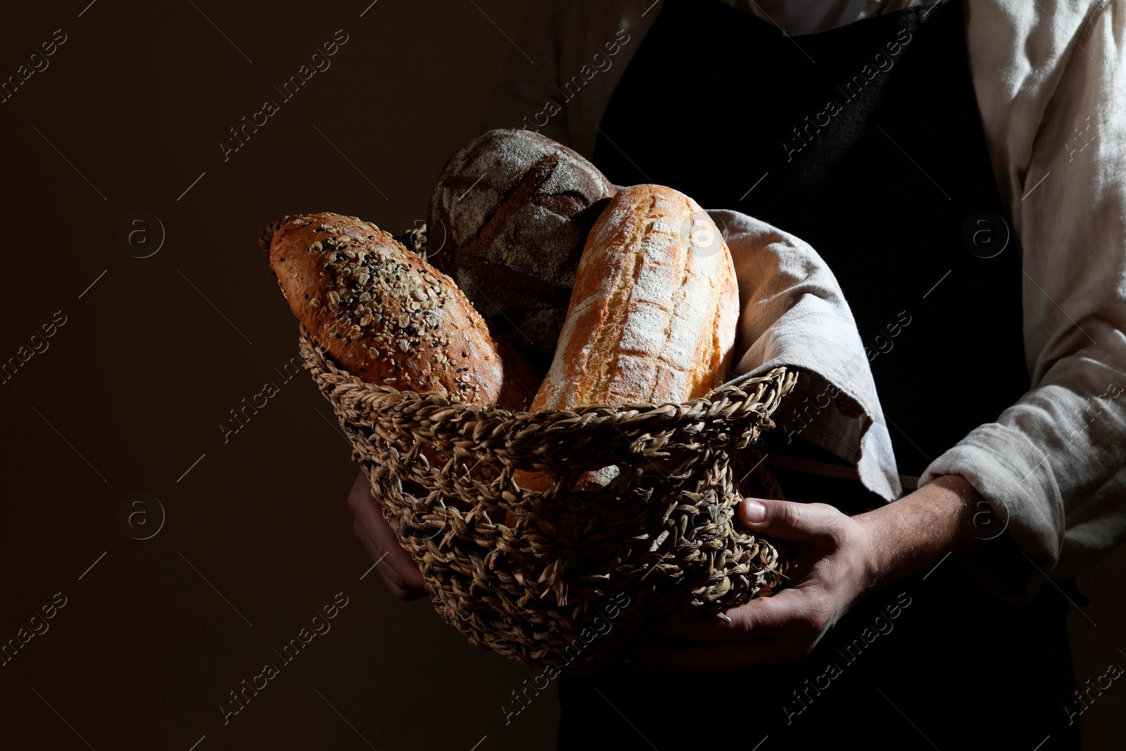 Photo of Man holding wicker basket with different types of bread on dark brown background, closeup