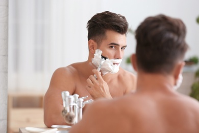 Photo of Young man shaving near mirror in bathroom