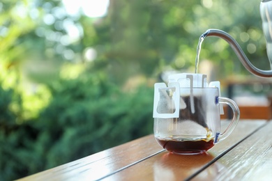 Photo of Pouring hot water into glass cup with drip coffee bag from kettle on wooden table outdoors. Space for text