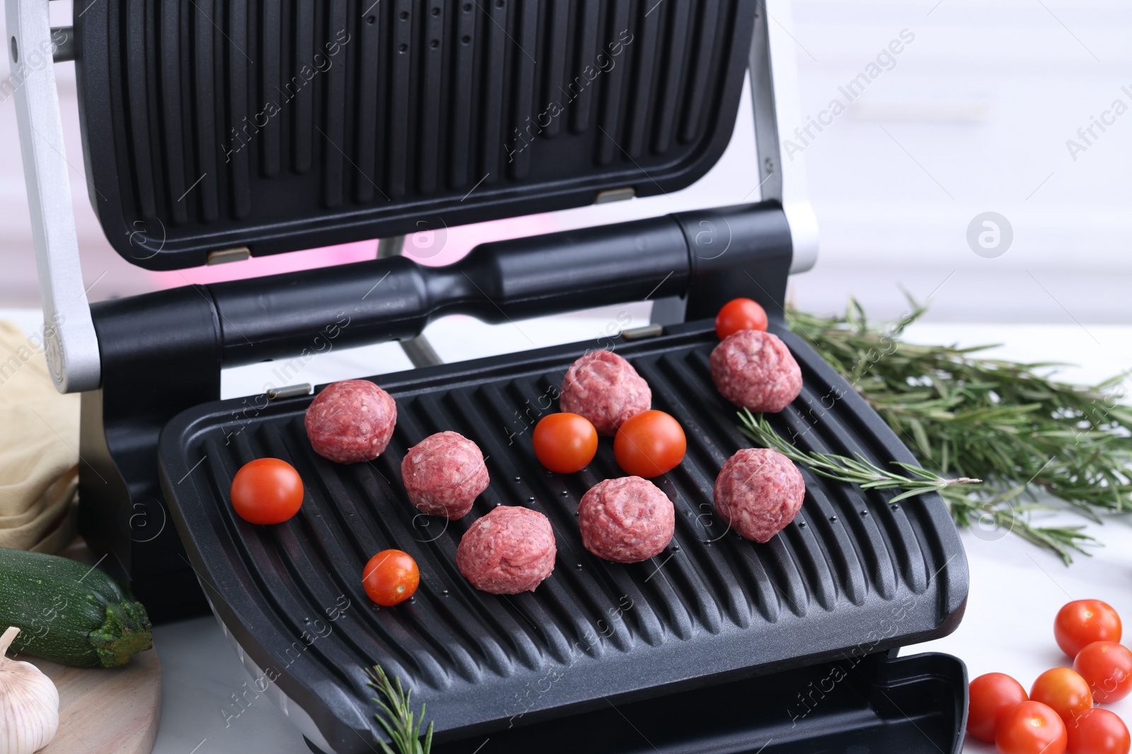 Photo of Electric grill with meatballs, tomatoes and rosemary on white table