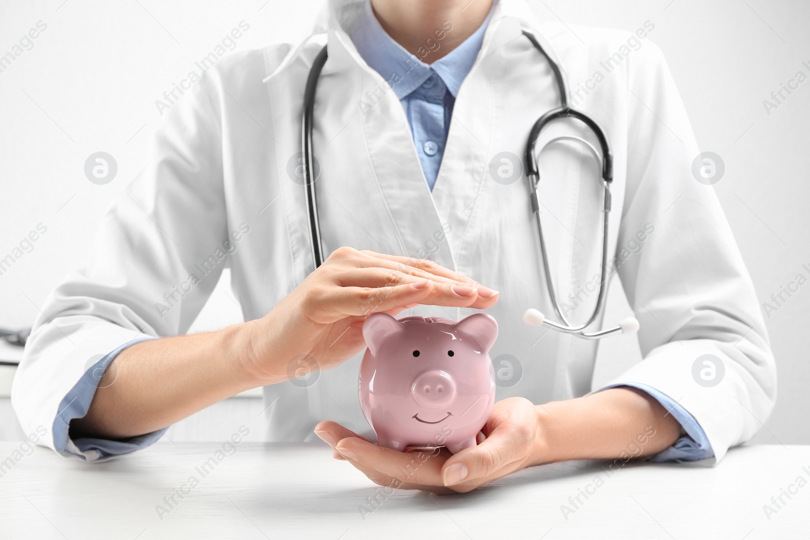 Photo of Doctor with piggybank at white table indoors, closeup. Medical insurance concept