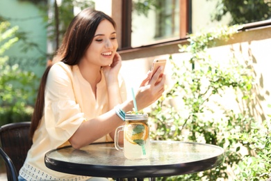 Photo of Young woman using mobile phone while drinking tasty lemonade at table, outdoors. Natural detox drink