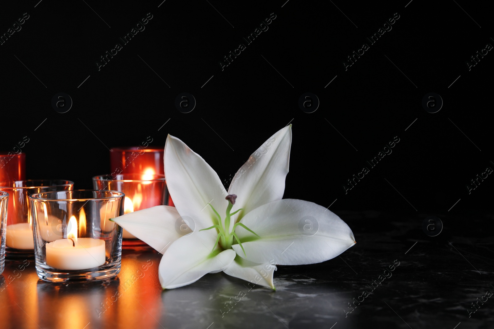 Photo of White lily and burning candles on table in darkness, space for text. Funeral symbol