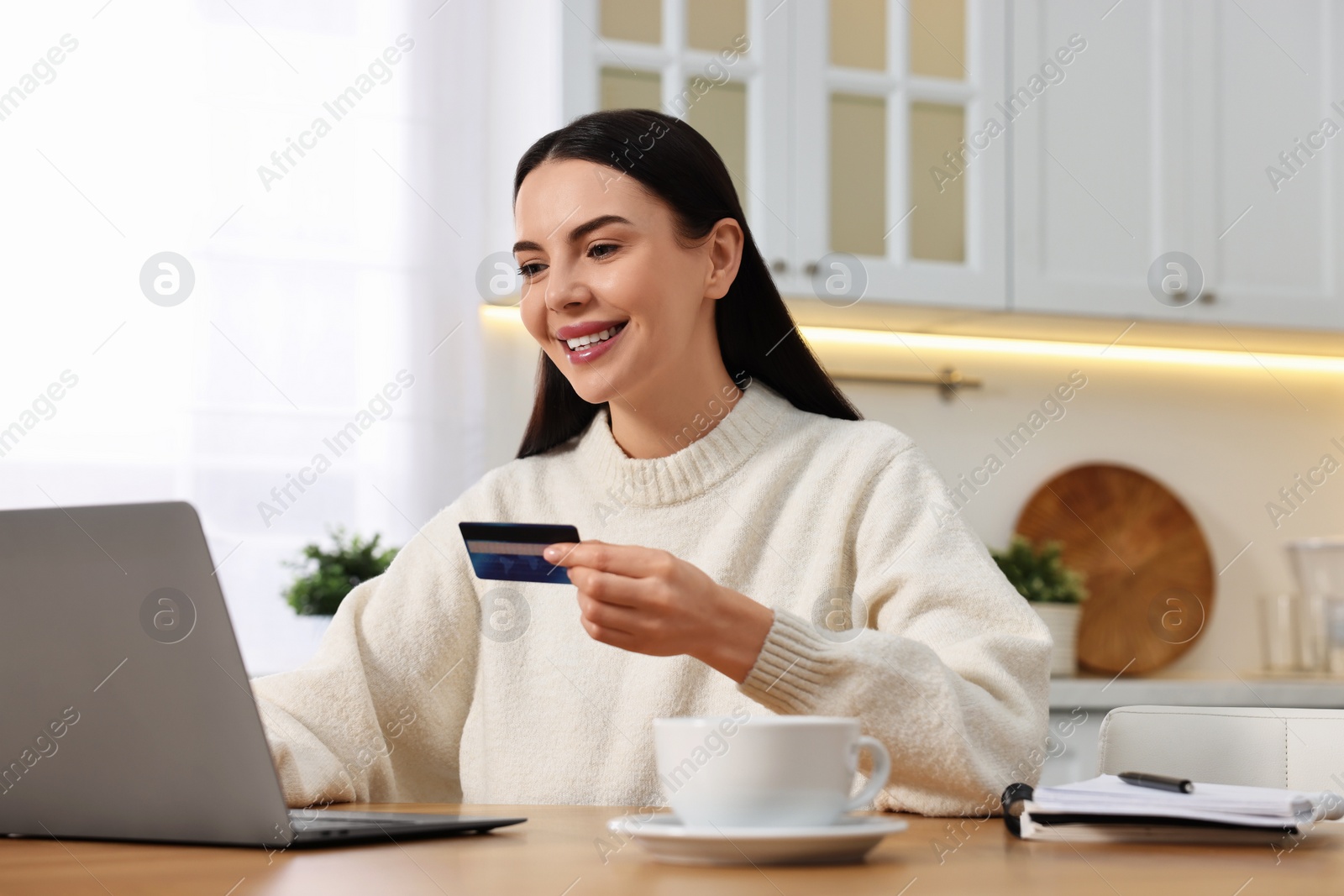 Photo of Happy young woman with credit card using laptop for shopping online at wooden table in kitchen