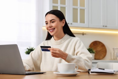 Happy young woman with credit card using laptop for shopping online at wooden table in kitchen