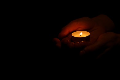 Woman holding burning candle in hands on black background, closeup. Space for text