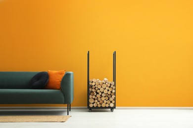 Stylish sofa with cushions, rug and firewood near orange wall indoors, space for text. Interior design