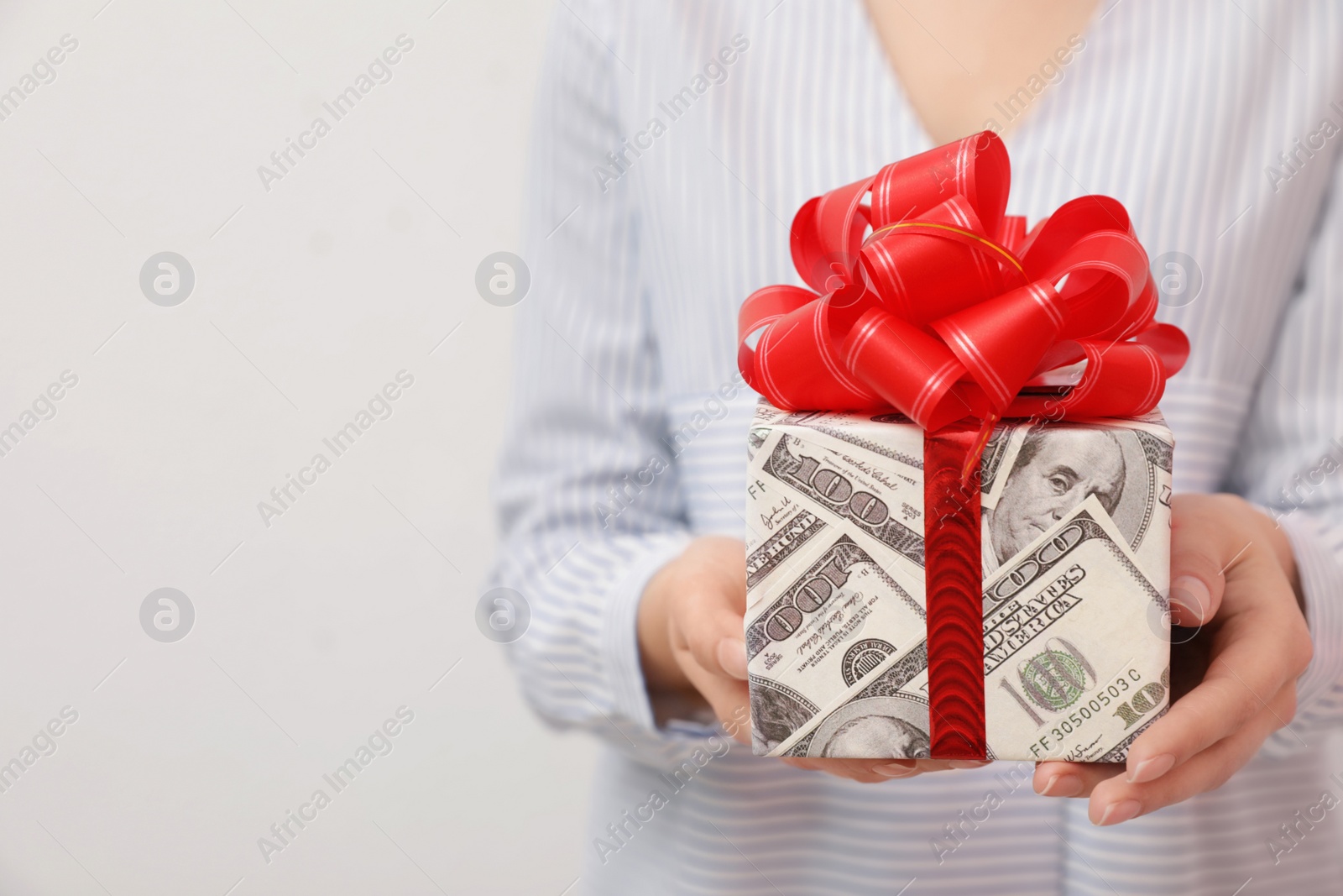 Photo of Woman holding gift box wrapped in dollars on light background, closeup