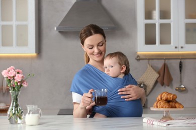 Mother with cup of drink holding her child in sling (baby carrier) in kitchen