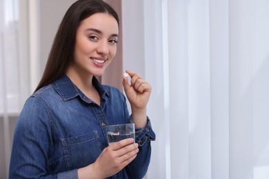 Photo of Beautiful young woman with glass of water and pill indoors, space for text
