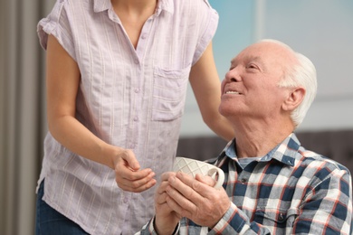 Photo of Elderly man with cup of tea near female caregiver at home
