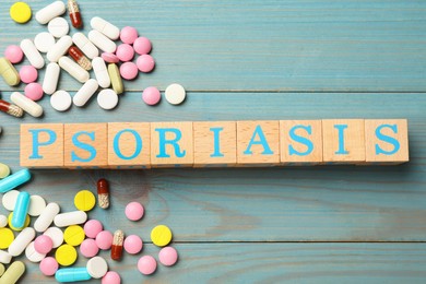 Photo of Word Psoriasis made of cubes with letters and pills on light blue wooden table, flat lay