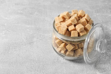 Photo of Brown sugar cubes in glass jar on light grey table, space for text