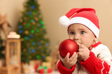 Photo of Cute little baby in sweater and Santa hat with Christmas bauble at home, space for text. Winter holiday