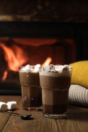 Photo of Glasses with hot cocoa, marshmallows and sweaters on wooden table near fireplace