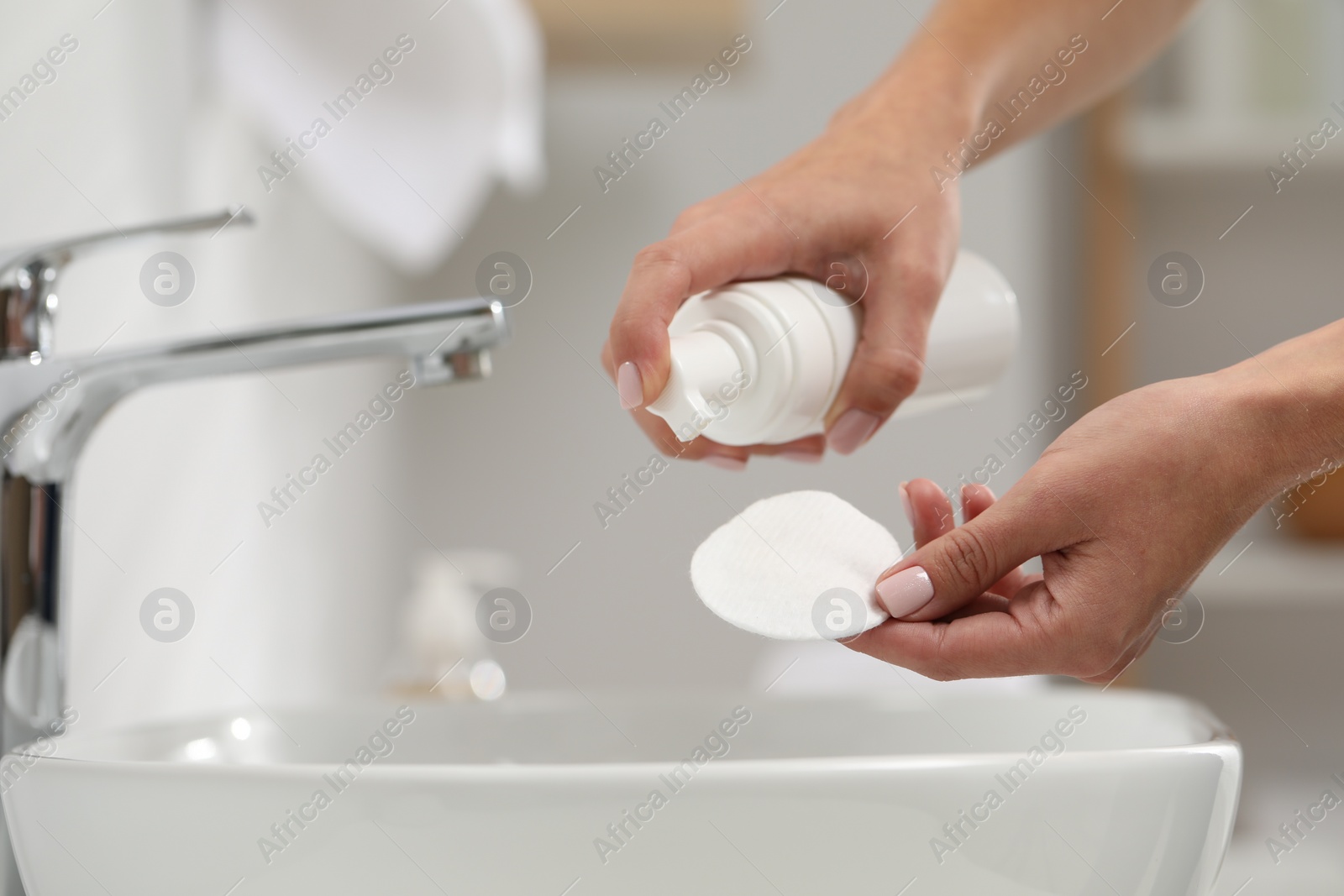 Photo of Woman applying makeup remover from bottle onto cotton pad in bathroom, closeup