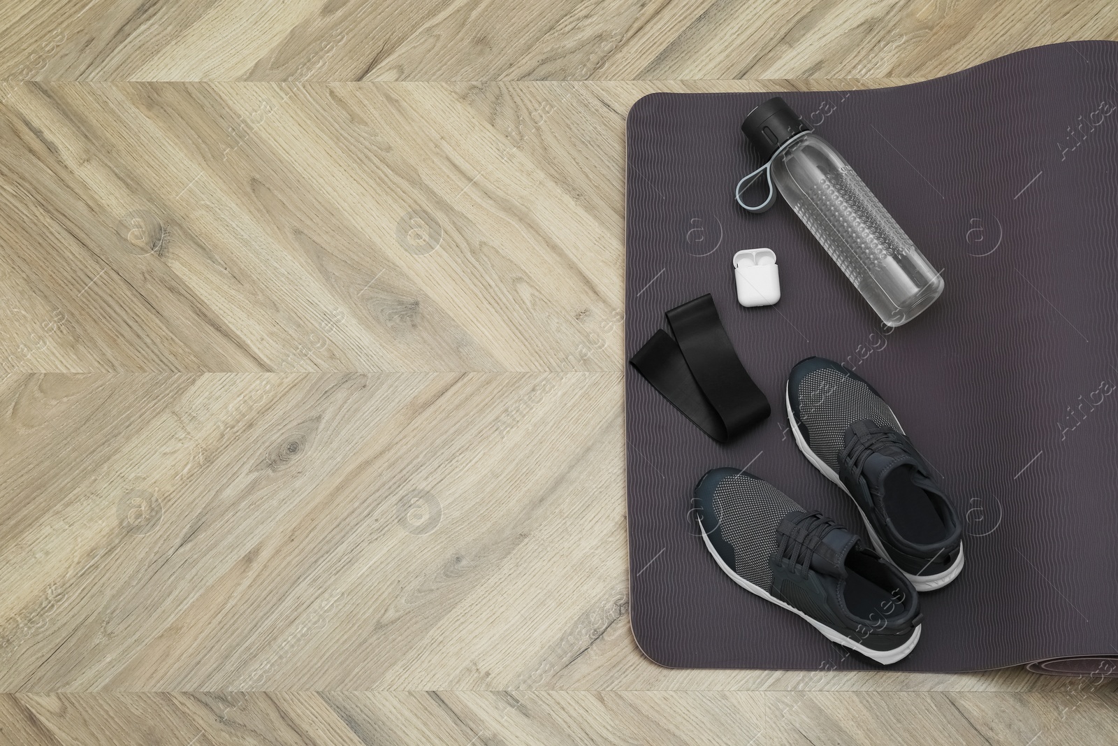 Photo of Exercise mat, bottle of water, wireless earphones, fitness elastic band and shoes on wooden floor, top view. Space for text