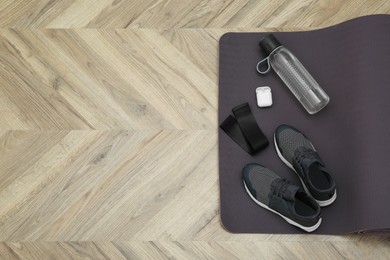 Photo of Exercise mat, bottle of water, wireless earphones, fitness elastic band and shoes on wooden floor, top view. Space for text