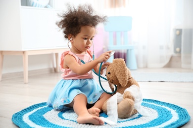 Photo of Cute African American child imagining herself as doctor while playing with stethoscope and toy bunny at home