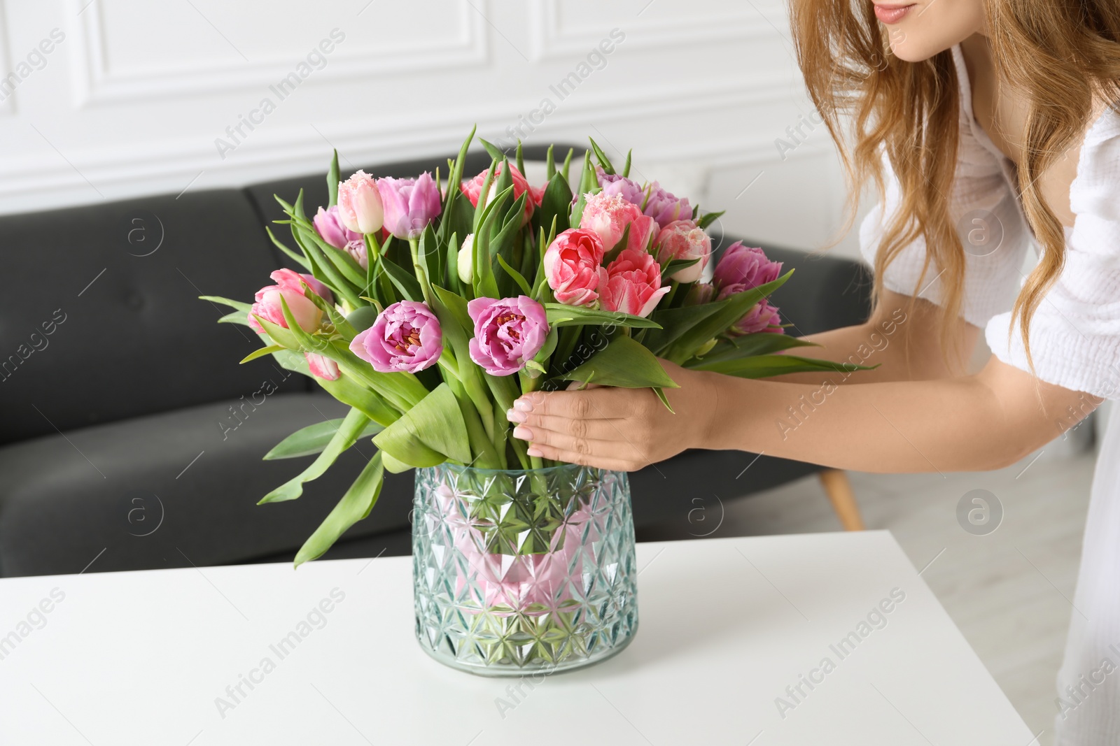 Photo of Woman putting bouquet of beautiful tulips in vase on white table indoors, closeup