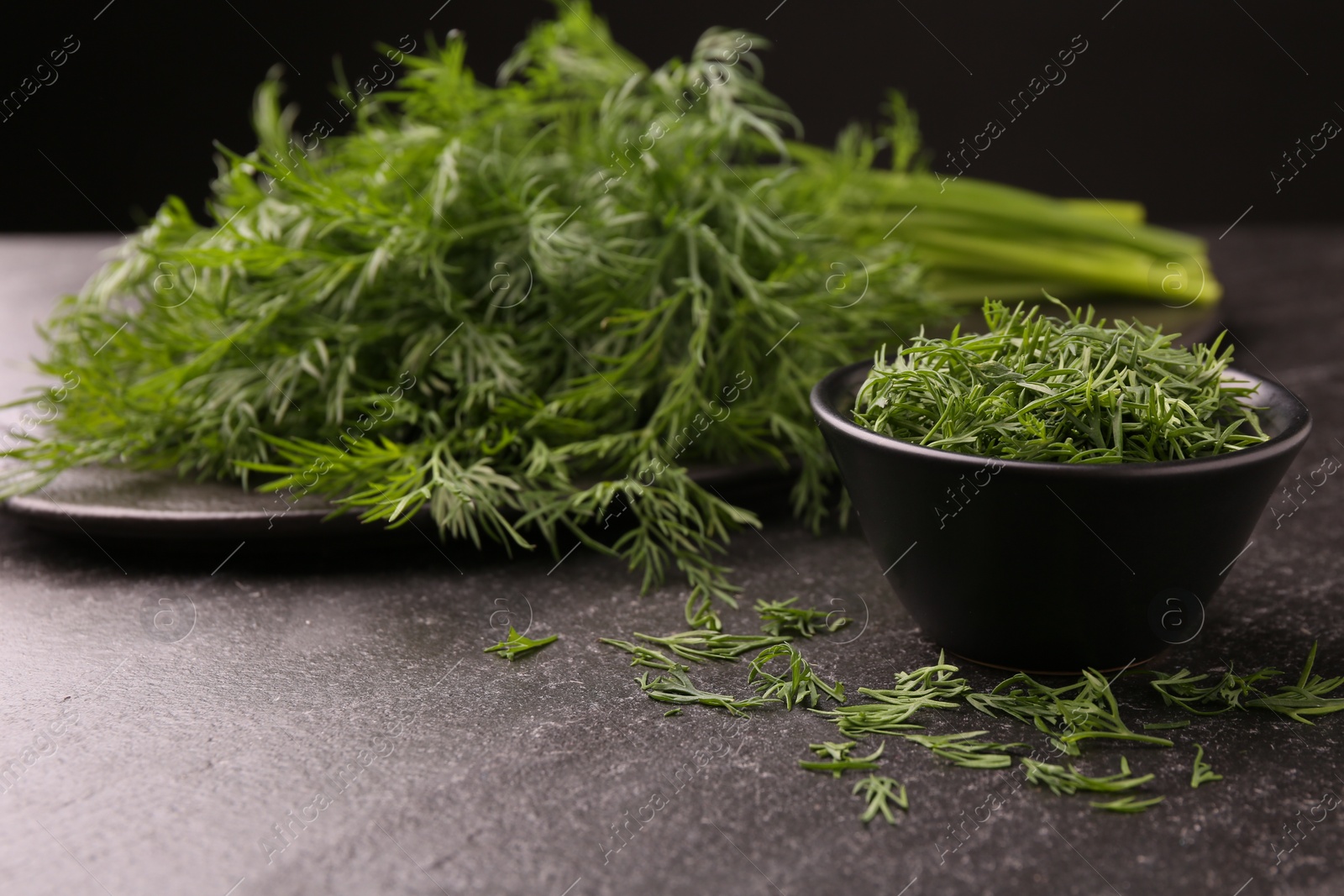 Photo of Sprigs of fresh dill and bowl with cut one on dark textured table, closeup