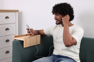 Happy man holding smartphone and listening to music on sofa with wooden armrest table at home