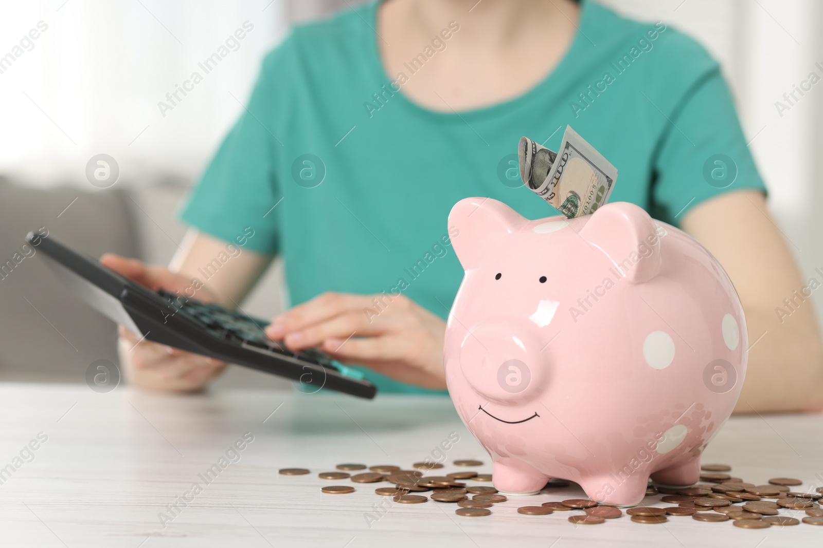 Photo of Financial savings. Woman using calculator at white wooden table indoors, focus on piggy bank and coins