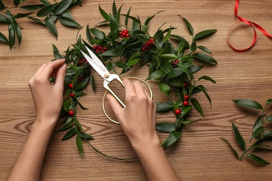 Photo of Florist making beautiful mistletoe wreath at wooden table, top view. Traditional Christmas decor