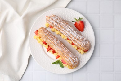 Delicious eclairs filled with cream, strawberries and mint on white tiled table, top view