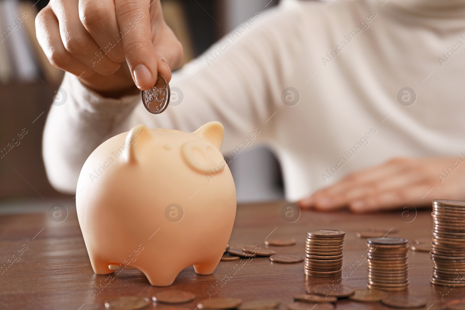 Photo of Woman putting coin into piggy bank at wooden table indoors, closeup