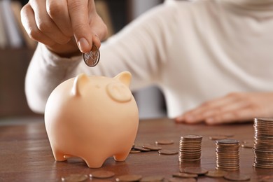 Photo of Woman putting coin into piggy bank at wooden table indoors, closeup